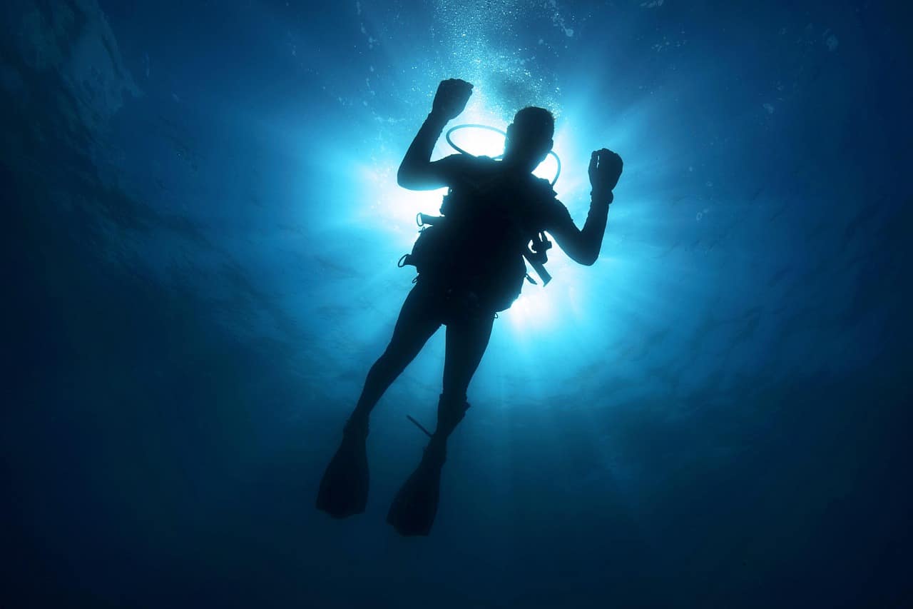 A diver in silhouette mid-frame in deep cobalt water, with shafts of surface light cutting down